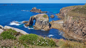 Enys Dodnan Longships This landscape photograph shows the Enys Dodnan Arch and the surrounding cliffs on the coast of Cornwall, England, in the United Kingdom. The image was captured close to midday in summer, with the sunlight illuminating the blue sea and highlighting the rocky formations. The Enys Dodnan Arch, a natural rock structure, is prominently situated near the cliffs, and beyond it, the Longships Lighthouse can be seen on the horizon. Coastal vegetation and wildflowers are visible in the foreground, enhancing the view of the rugged shoreline in this region of Cornwall.